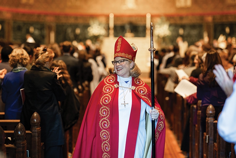 Woman wearing red bishop garments and mitre, walking down church aisle