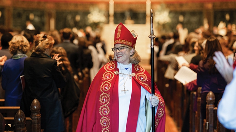 Woman wearing red bishop garments and mitre, walking down church aisle