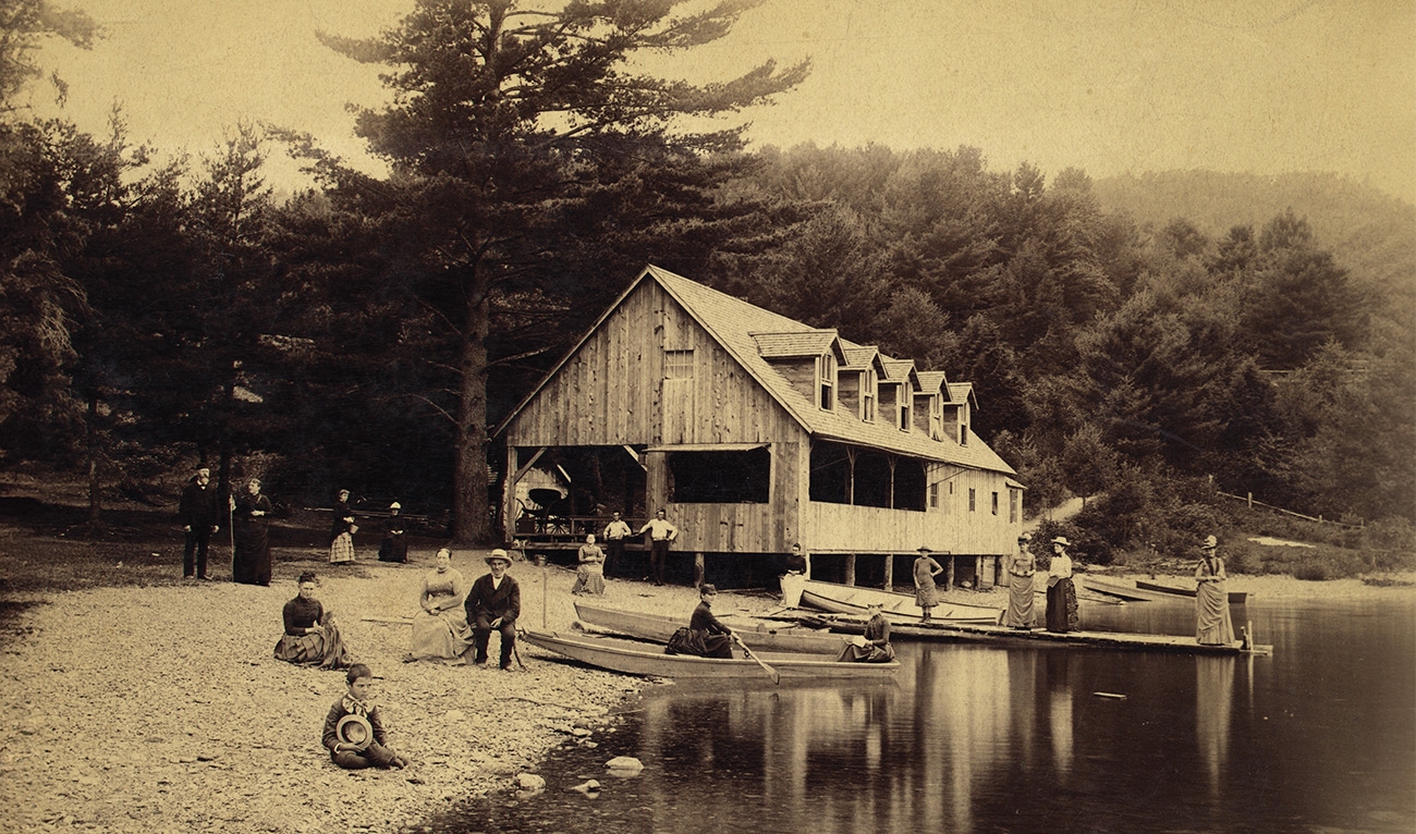 Historic, sepia-toned photo of people at the water's edge at a camp, some in boats