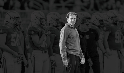 Football coach with headset on and players behind him