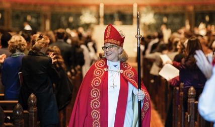 Woman wearing red bishop garments and mitre, walking down church aisle